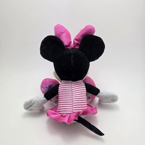 May include: A plush Minnie Mouse doll, viewed from the back, with black ears and a pink bow. The doll wears a pink and white striped dress with a matching skirt. Gray gloves and a black tail complete the design.