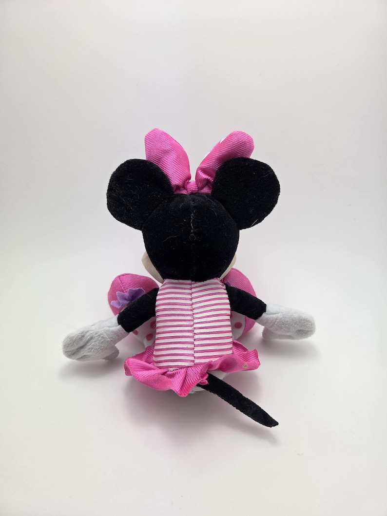 May include: A plush Minnie Mouse doll, viewed from the back, with black ears and a pink bow. The doll wears a pink and white striped dress with a matching skirt. Gray gloves and a black tail complete the design.