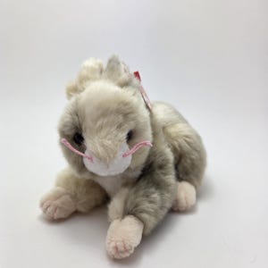 May include: A plush rabbit toy in shades of gray and white, with pink whiskers and paw pads. The stuffed animal is lying down on a white surface. A red tag is attached to the ear.