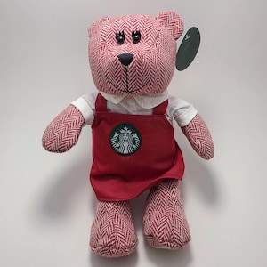 May include: A plush teddy bear featuring a red and white herringbone pattern fabric. The bear wears a white collared shirt underneath a red apron with a circular green and white Starbucks logo patch on the front. It has black plastic eyes and a tag.