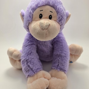 Op de afbeelding: Een pluche apenknuffel met zachte lavendelkleurige vacht op het hoofd, de armen en het lichaam, in contrast met lichtbeige stof op het gezicht, de oren, de handen en de voeten. De aap heeft geborduurde zwarte ogen en een kleine neus, zittend afgebeeld.