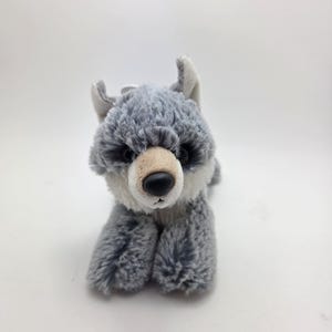 以下が含まれることがあります： グレー、白、黒の色合いのぬいぐるみオオカミのおもちゃ。 ぬいぐるみは柔らかく毛むくじゃらの質感で、薄いタン色の鼻と黒い鼻をしています。 オオカミは横たわっており、足を伸ばしています。