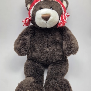 May include: A brown teddy bear wearing a red and white knitted hat. The bear has a white muzzle and foot pads. The hat has white trim and red tassels. The bear is standing upright against a white background.