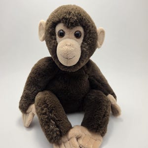 May include: A soft toy monkey with brown fur and a tan face, ears, hands, and feet. The plush animal has large, round eyes and is in a sitting position. The background is plain white.