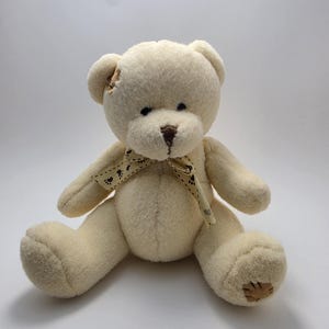 May include: A cream-colored plush teddy bear with a soft, fuzzy texture. The bear features a dark brown stitched nose, small black eyes, and a light-colored ribbon tied around its neck with a repeating dark brown paw print pattern.