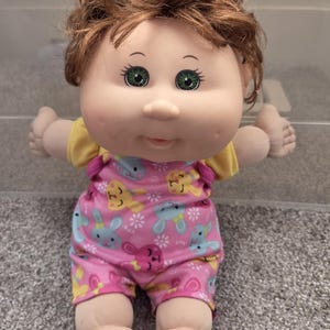 Peut inclure: Poupée vintage aux cheveux bruns et aux yeux verts. La poupée porte un haut jaune et une salopette rose avec un motif de lapin et de fleurs. La poupée a un corps doux et est assise.