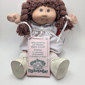 Peut inclure: Une poupée Cabbage Patch Kids avec des cheveux bruns en fil et une robe blanche. La poupée a un certificat de naissance et des papiers d'adoption roses. La poupée porte des chaussures et des chaussettes blanches.