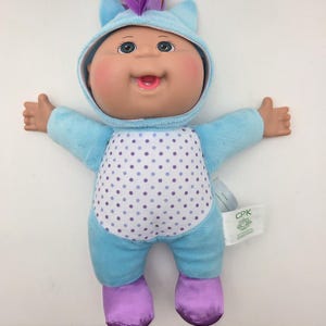 May include: A Cabbage Patch Kids doll wearing a light blue unicorn onesie with a white and purple polka-dot belly. The doll has a gold horn, purple ears, and purple feet. The doll's face is smiling.