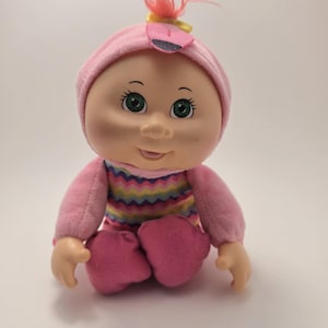 Pode incluir: Uma boneca de bebê vintage com um corpo macio e um rosto de vinil. A boneca usa um chapéu rosa com um detalhe de flor, uma camisa listrada e calças rosa. A boneca tem olhos verdes e uma expressão doce.