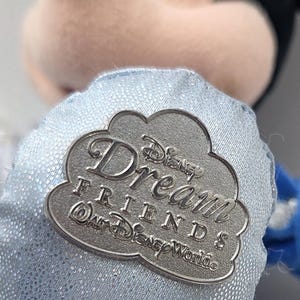 May include: A plush Mickey Mouse toy with a silver-colored pin. The pin is cloud-shaped and reads "Disney Dream Friends @ Walt Disney World". The toy has a light blue fabric with a glittery sheen.