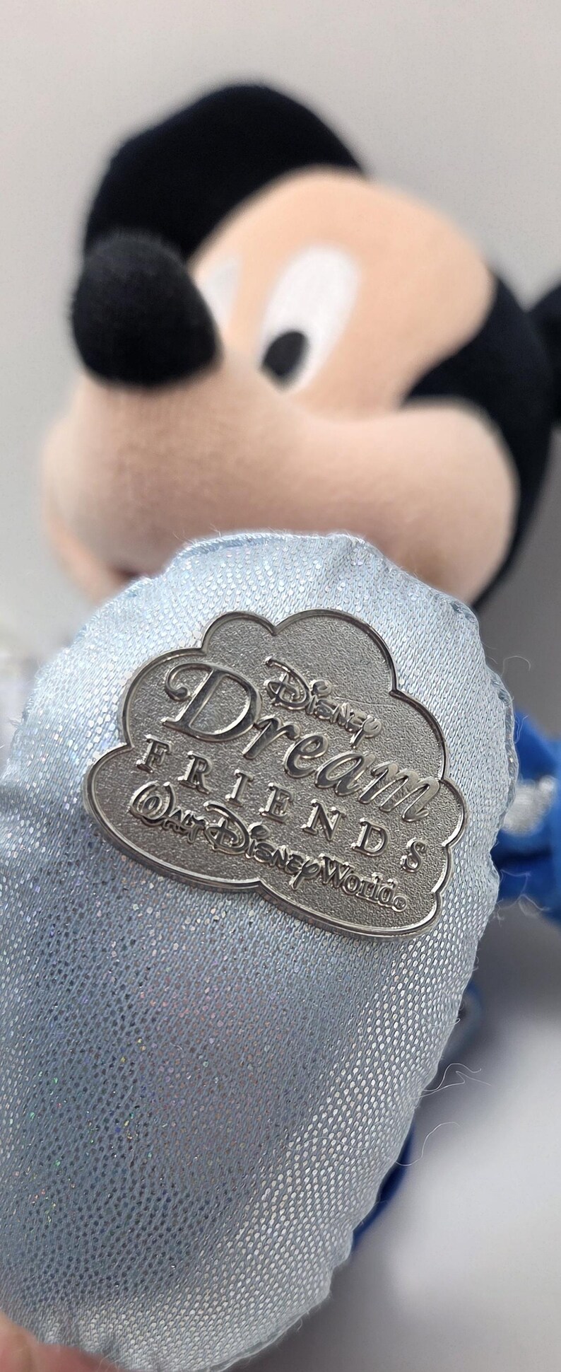 May include: A plush Mickey Mouse toy with a silver-colored pin. The pin is cloud-shaped and reads "Disney Dream Friends @ Walt Disney World". The toy has a light blue fabric with a glittery sheen.