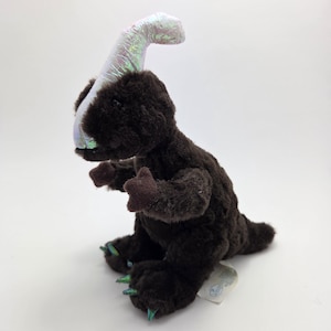 Puede incluir: Un juguete de peluche marrón de un dinosaurio Parasaurolophus con una cresta brillante e iridiscente. El juguete tiene brazos y pies pequeños de color marrón oscuro con garras verdes y azules. El dinosaurio está sentado sobre una superficie blanca.
