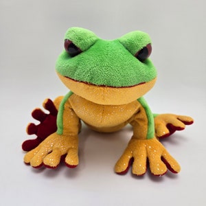 May include: Green and yellow plush frog with red feet. The frog has sparkly details on its body and large black eyes.