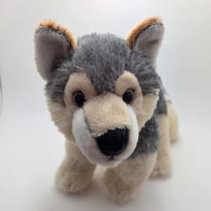 以下が含まれることがあります： リアルなデザインのぬいぐるみオオカミ。おもちゃは、グレーとタンの毛皮、黒い目、黒い鼻が特徴です。耳は茶色の先端があります。オオカミは四つん這いになっています。