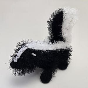 May include: A plush skunk toy with a black and white color scheme. The skunk has a fluffy, textured appearance, with a white stripe running down its back and a white patch on its face. The toy has small, black legs and a tail.