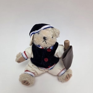 Puede incluir: Un oso de peluche que lleva una gorra de béisbol azul y blanca, una chaqueta azul marino con botones rojos y pantalones blancos con rayas azules y rojas. El oso lleva una bolsa de cuero marrón.