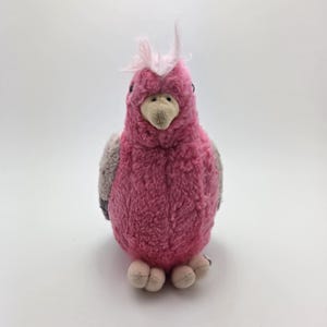 Può includere: Un peluche di un cacatua rosa e grigio con una cresta rosa chiaro. L'uccello ha un becco beige e piccoli piedi rotondi. Il giocattolo è realizzato in materiale morbido e strutturato ed è ambientato su uno sfondo bianco.