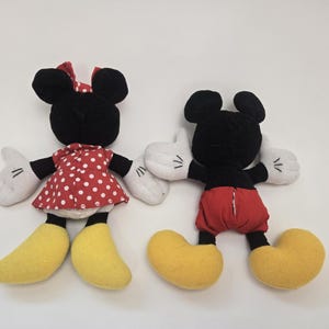 May include: A pair of plush toys, Mickey Mouse and Minnie Mouse, are shown from behind. Mickey is wearing red shorts and yellow shoes. Minnie is wearing a red dress with white polka dots and yellow shoes.