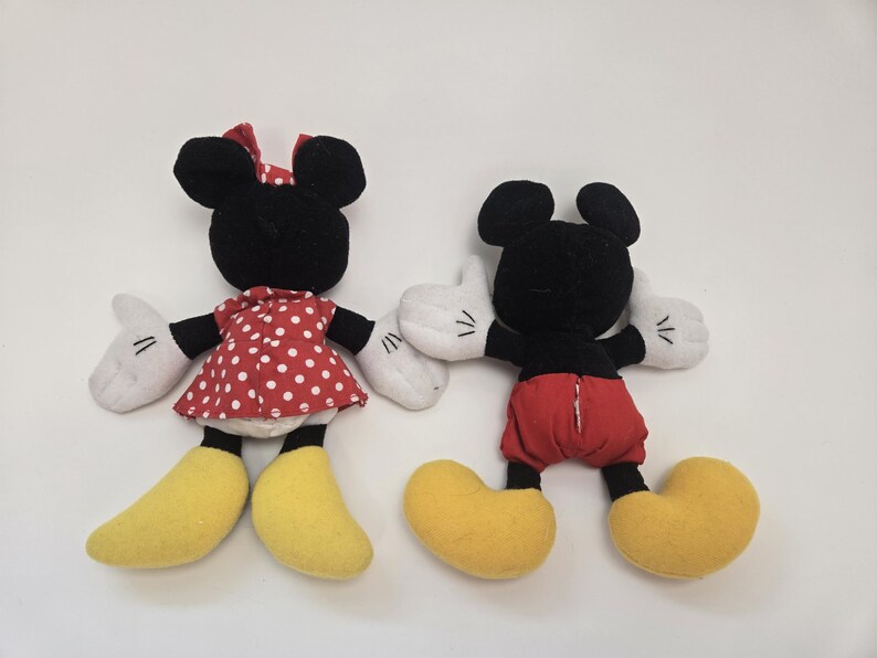 May include: A pair of plush toys, Mickey Mouse and Minnie Mouse, are shown from behind. Mickey is wearing red shorts and yellow shoes. Minnie is wearing a red dress with white polka dots and yellow shoes.