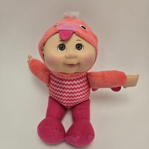 Könnte beinhalten: Rosafarbene Plüschpuppe Cabbage Patch Kid, die ein rosa-weißes Flamingo-Kostüm trägt. Die Puppe hat braune Augen, rosige Wangen und einen rosa Mund.