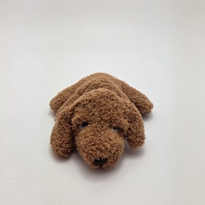 Puede incluir: Un juguete de peluche marrón con forma de perro y textura rizada. El juguete está tumbado, con la cabeza y las patas visibles. El perro tiene ojos negros y nariz negra. El fondo es blanco liso.