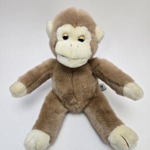 May include: A soft toy monkey with brown fur and cream-coloured facial features, hands, and feet. The stuffed animal has a smiling expression and is lying on a white surface. The monkey's arms and legs are outstretched.