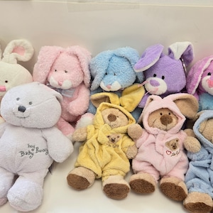 Op de afbeelding: Een verzameling pluche konijnen- en beren speelgoed in verschillende kleuren, waaronder roze, blauw, paars en wit. De speelgoedjes zijn allemaal zacht en knuffelig, met geborduurde details en sommige hebben de tekst "My Baby Bunny" erop.