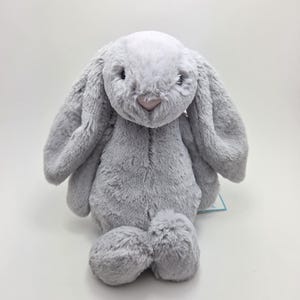 Puede incluir: Un suave conejito de peluche gris con orejas largas y caídas. El conejito tiene la nariz rosa y los ojos negros.