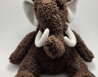 Könnte beinhalten: Ein braunes Plüsch-Wollhaarmammut mit texturiertem Fell, weißen Stoßzähnen und weißen Innenohren. Auf dem Kopf befindet sich ein Büschel längerer brauner Haare. Ein kleines rotes Ahornblatt ist auf den linken Fuß gestickt, roter Text befindet sich auf dem rechten Fuß.