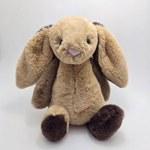 Peut inclure: Un lapin en peluche brun et beige avec des oreilles tombantes et des pieds bruns. Le lapin est assis les pieds joints.