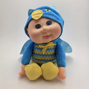 May include: A Cabbage Patch Kid doll wearing a blue owl costume with yellow accents. The doll has blue eyes, rosy cheeks, and a smiling expression. The costume includes a hood, wings, and a yellow bow.