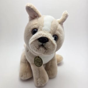 May include: A plush French Bulldog toy in tan and white. The stuffed animal has a white stripe on its head, black eyes, and a small gold tag that reads "My Pet Pals". The toy is sitting upright.
