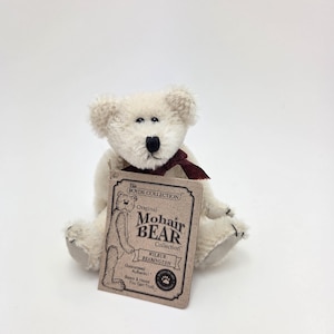 May include: A white mohair teddy bear with a burgundy ribbon around its neck. The bear is sitting on a white surface and has a tag that reads "The Boyds Collection Original Mohair Bear Collection Wilbur Bearington Guaranteed Authentic! Bears & Hanes You Can Trust."