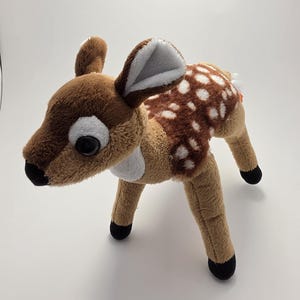 May include: A plush deer toy featuring soft brown fur, a white chest patch, and white spots on its back. The toy has large black eyes, a black nose, and black hooves. The ears are brown on the outside and white on the inside, standing upright.