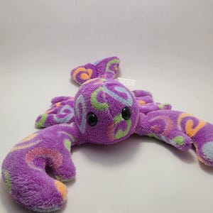 May include: A plush purple octopus toy with black button eyes and soft, curved tentacles. The octopus is decorated with colorful swirls of green, orange, and blue. The toy is designed for children.