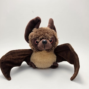 May include: A plush bat toy featuring dark brown wings and ears, a fuzzy medium brown head, and a light tan belly. The stuffed animal has a small black nose and round red eyes, with one ear standing upright and the other folded slightly downward.