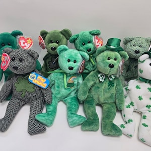 May include: A collection of nine green and white plush beanie baby bears. The bears are wearing shamrock and clover patterns and some have hats. The bears are all in a row on a white background.