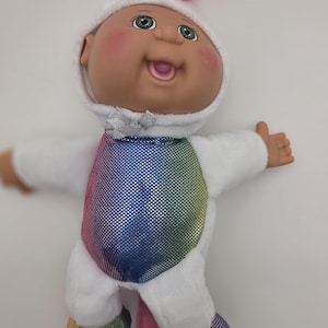 May include: A Cabbage Patch Kids doll dressed as a unicorn. The doll has a white hooded outfit with a rainbow-colored torso and matching feet. A pink horn and mane complete the unicorn look. The doll has green eyes and rosy cheeks.