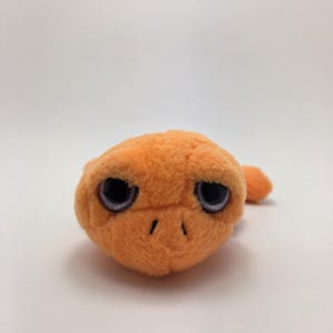 May include: An orange plush toy with large, black-rimmed eyes and a small, pointed tail. The toy has a soft, fuzzy texture and a simple design, resembling a small creature. The background is plain white.