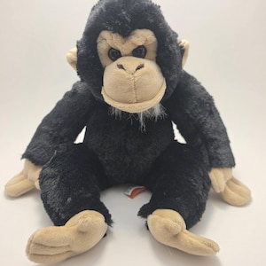 Può includere: Un peluche di scimpanzé con pelliccia nera e viso, mani e piedi color beige. L'animale di peluche ha un'espressione amichevole ed è seduto in posizione eretta. Il giocattolo è progettato per assomigliare a uno scimpanzé.