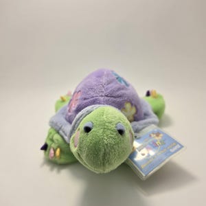 Peut inclure: Une peluche en forme de tortue avec une tête et des membres verts, une carapace lavande ornée d'applications florales et un ventre bleu-gris. Le jouet a une étiquette attachée. La tortue est une peluche.