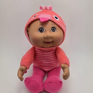 May include: A Cabbage Patch Kids doll wearing a pink flamingo costume. The doll has brown eyes and a smiling expression. The costume includes a hood with a beak, a zig-zag patterned shirt, and solid pink pants.