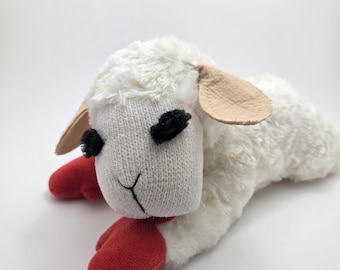 May include: A plush white lamb toy with a soft, textured body and a knitted face. The lamb has black stitched eyes, tan ears, and red feet. The toy is lying down on a white surface.