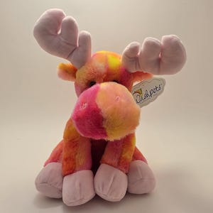 May include: A plush moose toy featuring a vibrant tie-dye pattern in shades of pink, orange, and yellow. The stuffed animal has soft, light pink antlers and hooves, with a tag attached to the ear that reads Wishpets A Wish Come True.