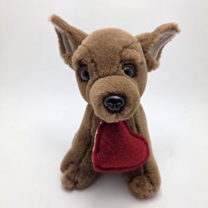 May include: A brown plush Chihuahua dog with large, expressive eyes and a black nose. The stuffed animal is holding a red heart with white stitching. The dog is sitting upright, with its ears perked up.