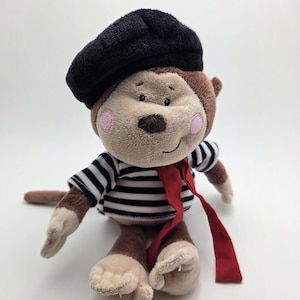 May include: A soft toy monkey dressed in a black beret, a black and white striped top, and a red scarf. The monkey has brown fur, beige hands and feet, and pink cheeks. The toy is designed to resemble a French stereotype.