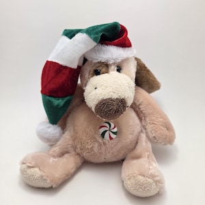 May include: A plush dog toy in a seated position, wearing a Christmas hat with red, white, and green stripes. The dog has a brown nose and ears, and a candy-shaped ornament is attached to its chest.
