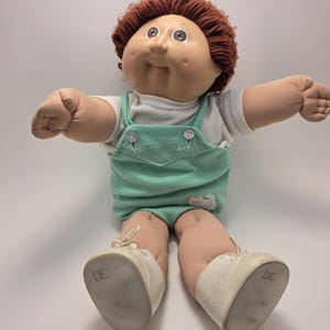 May include: A vintage Cabbage Patch Kid doll with a soft, vinyl face, brown yarn hair, and a mint green overall. The doll is wearing a white shirt and white shoes. The doll is sitting with its arms outstretched.