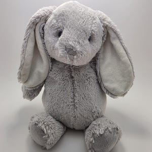 May include: A plush stuffed animal in the shape of a rabbit with soft, light gray fur. The toy features long, floppy ears, a rounded body, and a small stitched nose. The texture of the fur appears shaggy and dense across the entire surface.
