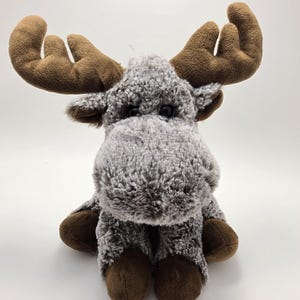 May include: A plush moose toy featuring a textured grey and white mottled fur body, large brown antlers, and dark brown hooves. The stuffed animal has two small black eyes and a prominent, rounded snout, sitting in an upright position.